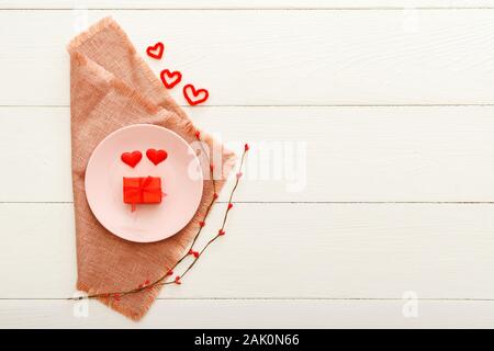 Valentines Day table setting on a white wooden table. Dinner meal in restaurant at Valentine Day.Two red hearts and present gift on pink plate Stock Photo
