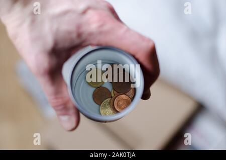A person holding a cup with some coins begging for more change. Stock Photo