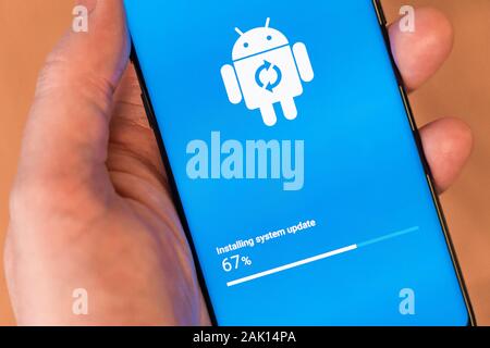 A man's hand holding a smartphone installing an Android system update, showing the Google Android mascot robot - Bugdroid Stock Photo