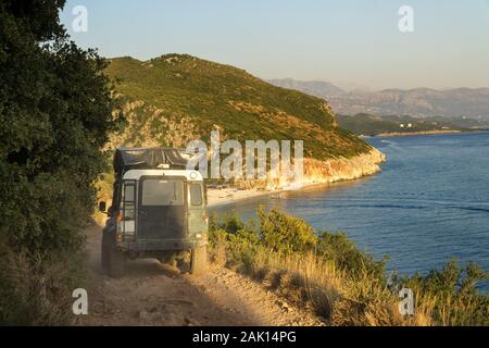 4x4 off-road car with tent on the roof travels along a dirt road above the precipice towards the beach of Gjipe gorge in Albania in summer at sunset. Stock Photo