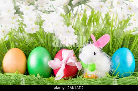 Easter Colorful Decorated Eggs and Little Toy Bunny on Green Grass Background With White Flowers Easter Card. Stock Photo