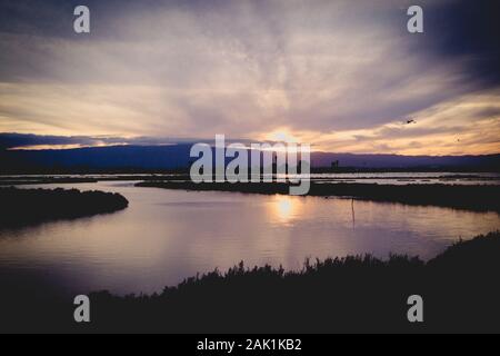 Sun setting behind the mountains, behind snaking river on a cloudy winter's day. Purple haze, a reflection of the sky. Stock Photo