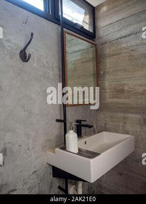 Bathroom modern loft style. White bathroom sink with faucet with liquid soap bottle and mirror and cloth hanger on concrete wall, vertical style. Stock Photo