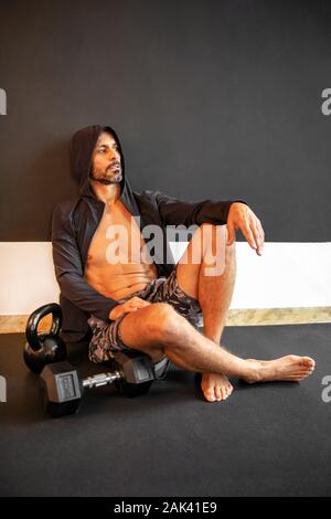 Bearded man in hoodie resting up after a workout sitting on the floor of the gym relaxing alongside kettlebell weights in a healthy active lifestyle c Stock Photo