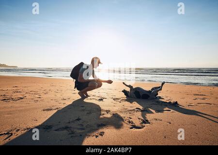 Young man playing with cheerful dog on sand beach against sea, Sri Lanka. Stock Photo