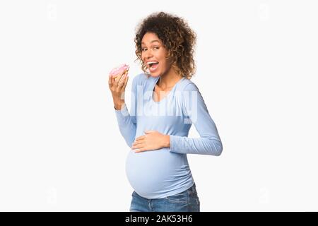 Hungry expectant woman eating donut, standing over white studio background, free space Stock Photo