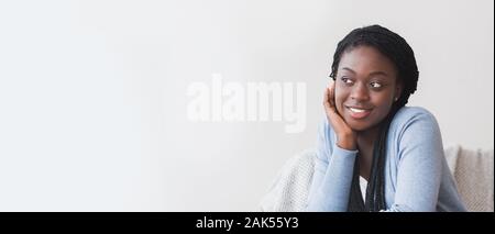 Morning relax. Pensive afro woman resting her head on hand, sitting on couch at home and looking aside at copy space, panorama Stock Photo