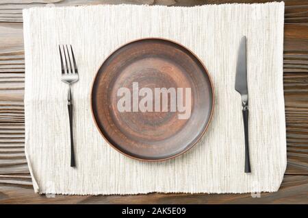 Top view at empty clay plate knife and fork aside on grey napkin on brown wooden kitchen table Stock Photo
