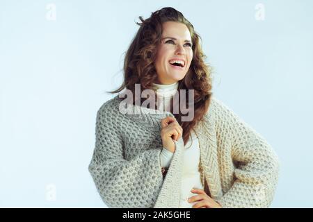 happy modern housewife with long wavy hair in neck sweater and cardigan looking at copy space against winter light blue background. Stock Photo