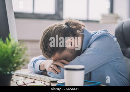Fell asleep. Young bearded man in blue shirt having a nap at working place Stock Photo