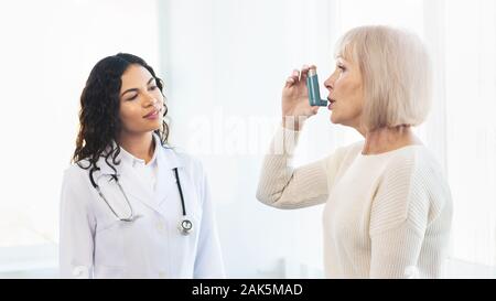 Medical Advice. Elderly woman using asthma inhaler, mexican doctor watching, free space, panorama Stock Photo