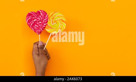Black woman's hand holding two spiral lollipops over orange background, panorama with free space Stock Photo