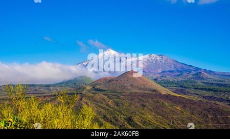 Spain, Tenerife, Colorful plant covered mountains and volcanoes in teide national park, covered by white snow and fast moving fog clouds on sunny day Stock Photo