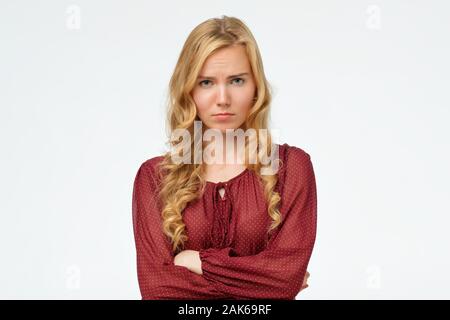 Beautiful girl with blonde long hair frowning her face in displeasure, keeping arms folded. Attractive young woman in closed posture. Studio shot Stock Photo