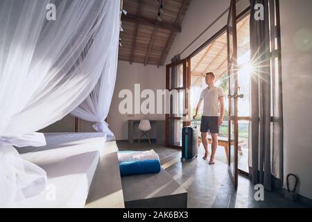 Luxury accommodation in tropical destination. Young man with suitcase entering to modern bungalow. Stock Photo