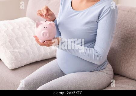 Pregnancy And Budget. Unrecognizable Pregnant Lady Putting Coin In Piggybank Sitting On Sofa At Home. Cropped Stock Photo