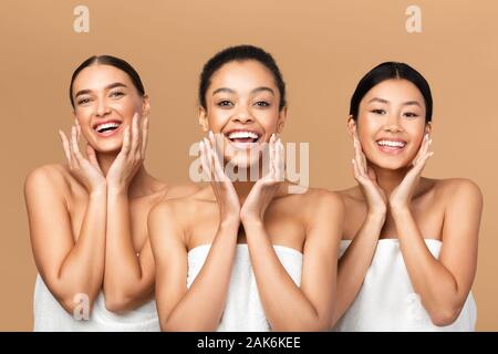 Skincare And Beauty. Three Multiracial Girls Touching Face With Perfect Smooth Skin On Cheeks Smiling At Camera Over Beige Background Stock Photo