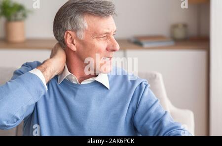 Senior Man Massaging Neck Suffering From Arthritis Pain Sitting On Couch At Home. Selective Focus Stock Photo