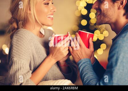 Cute couple enjoying hot tea on Christmas eve, talking to each other against xmas tree Stock Photo