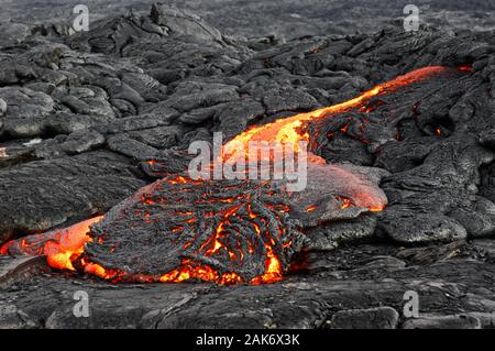Hot magma of an active lava flow emerges from a fissure and flows over previously deposited dark, strongly structured rock, the glowing lava shows its Stock Photo
