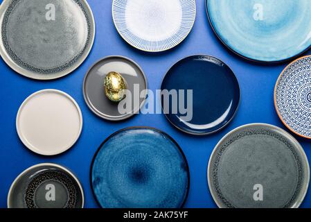 Different Ceramic Plates with One Easter Egg of pastel colors on blue background. Happy Easter card concept, minimalistic design, top view Stock Photo
