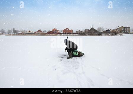 A fisherman sits near an ice hole on a frozen snow-covered river in a snowfall Stock Photo