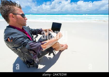 Castaway survivor businessman sitting on a tropical beach in a ragged suit watching his tablet computer Stock Photo