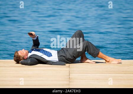Relaxed barefoot businessman lying on a sunny dock by the sea using his smartphone Stock Photo