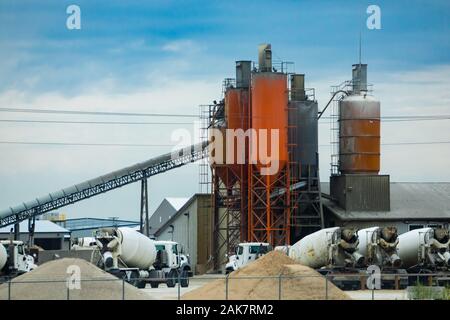 A wide angle view of a concrete batching and manufacture plant with aggregate conveyor, cement silos, industrial architecture and parked mixer trucks Stock Photo