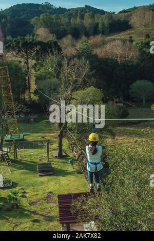 Girl descending by cable in a zip-line over meadows and trees on a valley near Bento Goncalves. A wine producing country city in southern Brazil. Stock Photo