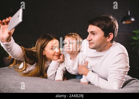 A young family lies on the bed and looks at the mobile phone. Mom, dad and son are watching a video on a smartphone in the bedroom in the evening Stock Photo