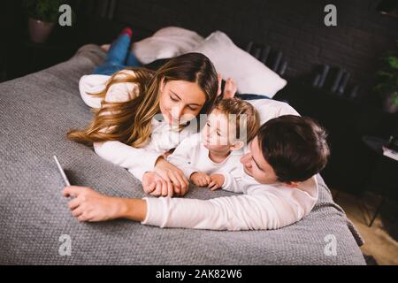 A young family lies on the bed and looks at the mobile phone. Mom, dad and son are watching a video on a smartphone in the bedroom in the evening Stock Photo