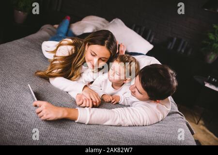 A young family lies on the bed and looks at the mobile phone. Mom, dad and son are watching a video on a smartphone in the bedroom in the evening Stock Photo