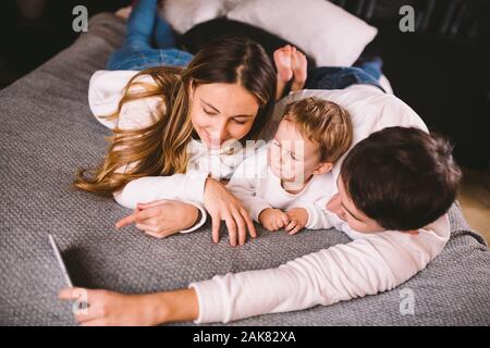 A young family lies on the bed and looks at the mobile phone. Mom, dad and son are watching a video on a smartphone in the bedroom in the evening Stock Photo