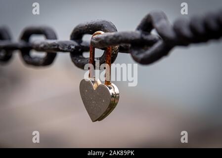 A romantic closed and rusty heart shaped padlock hanging from a chain beside a river with blurred cityscape in the background. Concept of long lasting Stock Photo