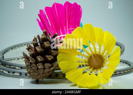 Badminton rackets and colorful, pink and yellow shuttlecocks of natural feathers placed on grey surface with pine cone and viewed in close-up. Amateur Stock Photo