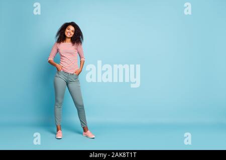 Full length photo of amazing pretty dark skin lady with adorable smile holding hands in pockets wear casual clothes isolated pastel blue color Stock Photo