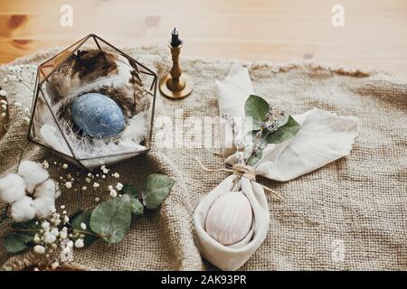 Stylish easter eggs, modern nest, napkin bunny ears on rustic table. Natural dyed easter eggs with eucalyptus branch, spring flowers, cotton on rural Stock Photo