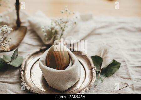 Rustic Easter table decorations. Stylish Easter brunch table setting with egg in easter bunny napkin. Modern natural dyed green egg on napkin with bun Stock Photo