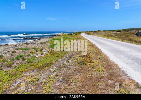On Robben Island a narrow road runs parallel to the rocky shorline and the blue water of Table Bay Stock Photo