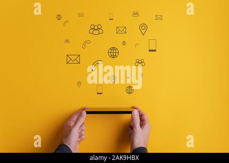 Male hands holding smart phone horizontally with contact and communication icons coming out of it in a conceptual image. Over yellow background. Stock Photo