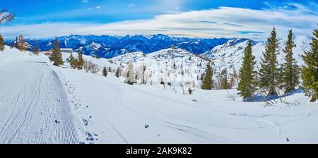 Winter panorama of Feuerkogel Mountain plateau with wide groomed piste, covered with corduroy pattern, snowy valley with coniferous trees and hazy roc Stock Photo