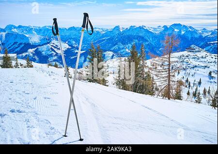 Alpine winter landscape from the Feuerkogel Mountain plateau with a view on crossed ski poles, groomed piste and blue Dachstein peaks on the backgroun Stock Photo