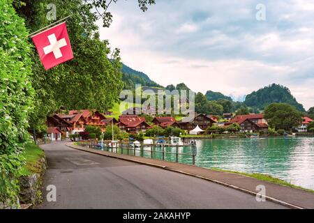 Swiss flag waving and traditional wood house and boats on the Lake Brienz in swiss village Iseltwald, Switzerland Stock Photo