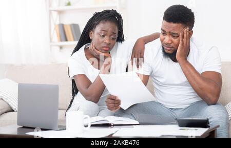 Debt concept. Portrait of sad afro couple looking at invoice, upset about huge bills, not able to pay mortgage, sitting depressed on sofa at home Stock Photo