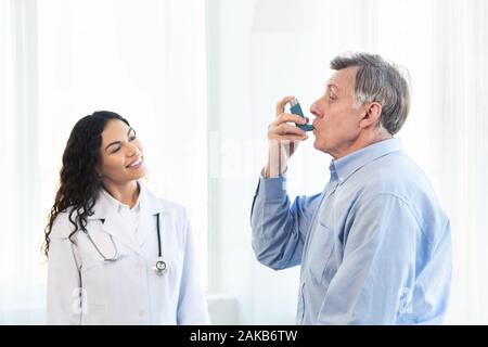 Medical Advice. Elderly man using asthma inhaler, mexican doctor watching, free space Stock Photo