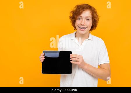 red-haired curly boy consultant in a store in a white T-shirt shows a tablet display with a layout on a yellow background Stock Photo