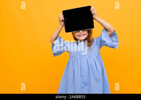charming attractive caucasian girl in a dress holds a tablet with a layout in front of her eyes on a yellow background