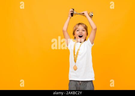 happy boy in a t-shirt with a medal on his neck raises the winner's cup on a yellow background with copy space Stock Photo