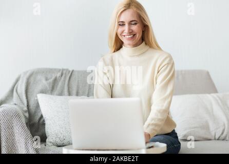 Excited middle-aged woman reading article online on laptop, relaxing on sofa at home Stock Photo